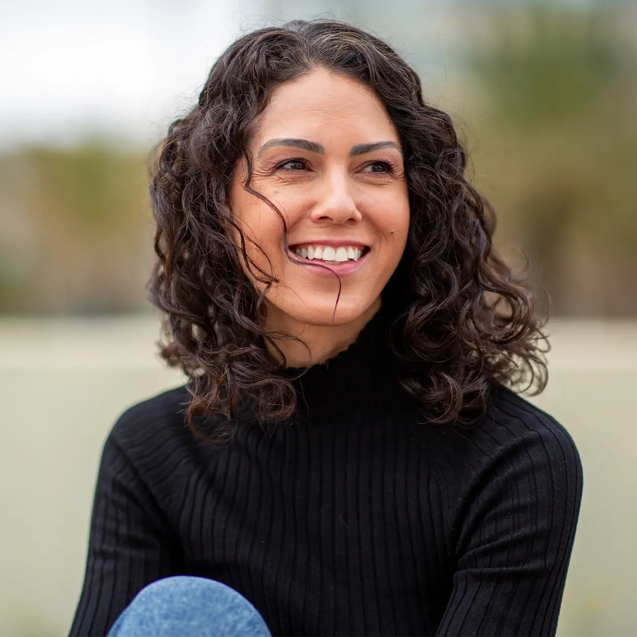 Smiling woman with curly hair in a natural outdoor setting.